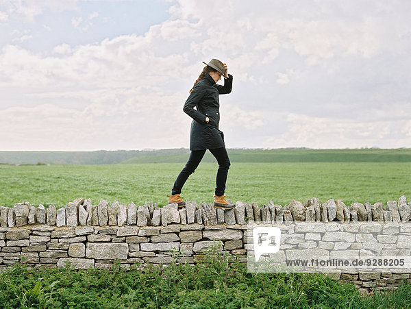 Eine Frau mit Hut  die auf einem Feld an der Spitze einer Trockenmauer entlanggeht.