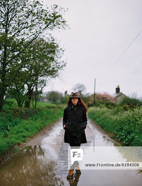 Eine Frau  die auf einer nassen Straße geht und einen Mantel und einen Hut trägt.