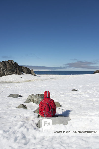 Eine Person in einer orangefarbenen Jacke sitzt und betrachtet die Landschaft auf einer antarktischen Insel.