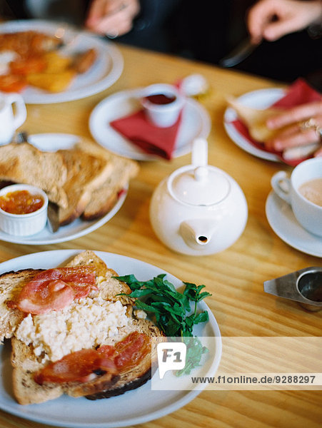 A table covered with plates of food. A cooked breakfast of bacon  eggs and toast and pot of tea. Two people seated