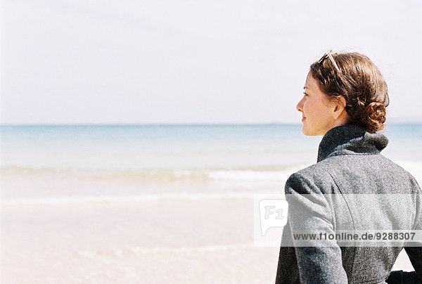 Eine Frau in einer grauen Jacke mit Blick auf den Strand und das Meer.