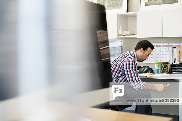 A man working in an office  sitting checking his smart phone.