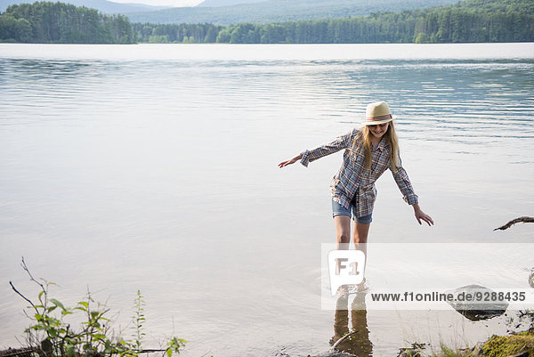A young girl in a straw hat and shorts paddling in the shallow waters of a lake.