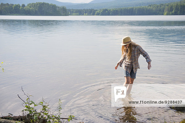 A young girl in a straw hat and shorts paddling in the shallow waters of a lake.