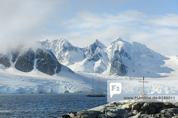 A cross and grave on a hilltop overlooking the snow capped peaks of the mountains on Petterman island. A tall masted ship in the channel.