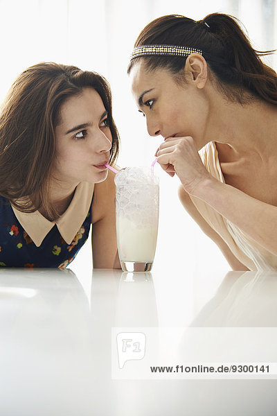 Mutter und Tochter blasen Blasen in Milch  während sie sich am Tisch ansehen.