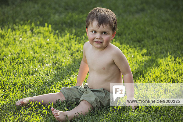 Volle Länge des süßen Jungen  der auf Gras im Park sitzt.