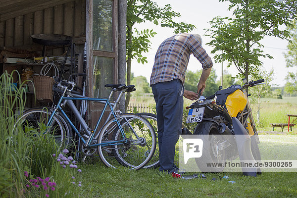 Rückansicht des reifen Mannes,  der das Motorrad auf dem Hof repariert.
