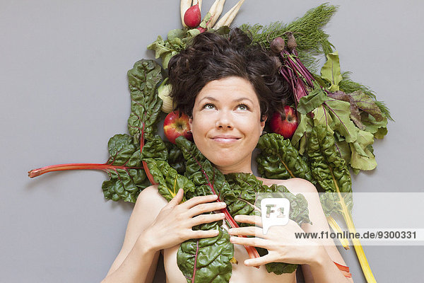 Eine Frau  die sich mit Rhabarberblättern bedeckt  während sie mit Gemüse auf grauem Hintergrund liegt.