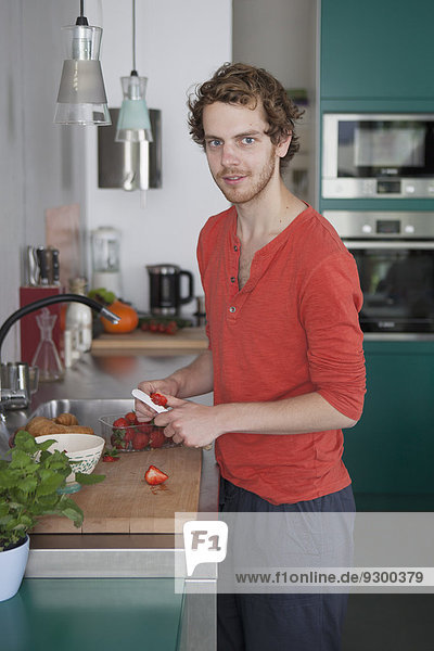 Porträt eines jungen Mannes  der Erdbeeren an der Küchentheke schneidet.