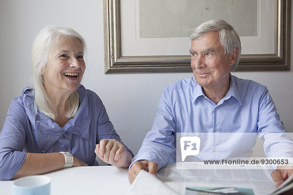 Happy senior couple with newspaper sitting at table in house