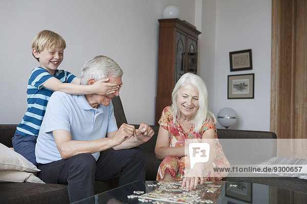 Junge  der die Augen des Großvaters bedeckt  während die ältere Frau ein Puzzle am Tisch im Wohnzimmer arrangiert.