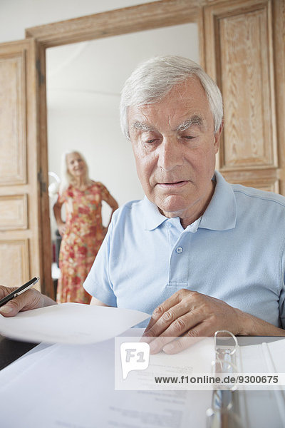 Senior-Mann bei der Prüfung von Finanzdokumenten mit Frau im Hintergrund zu Hause