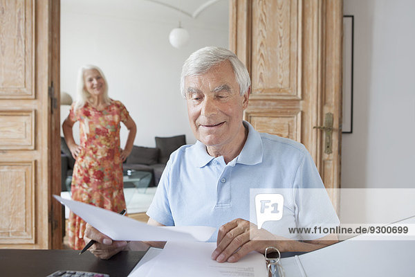 Senior-Mann  der zu Hause mit Frau im Hintergrund Finanzdokumente prüft