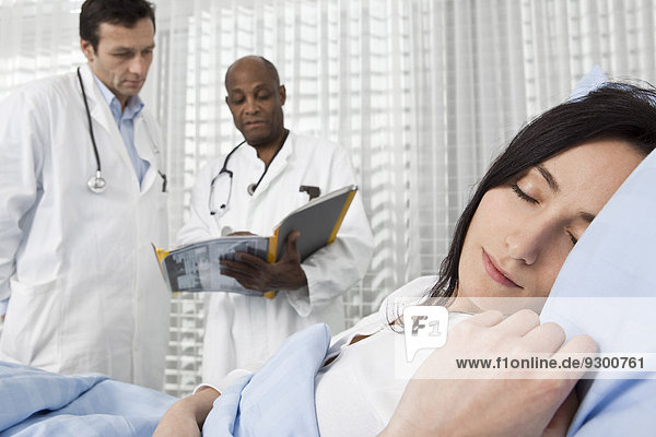 Eine Patientin schläft in einem Krankenhausbett  während zwei Ärzte im Hintergrund über ihre Akte sprechen.