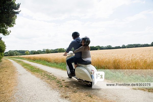 Rear view of mature man and daughter riding motor scooter along dirt track
