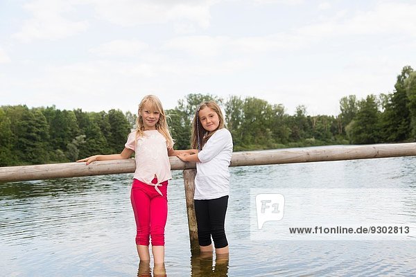 Portrait of two girls holding onto fence whilst paddling in rural lake