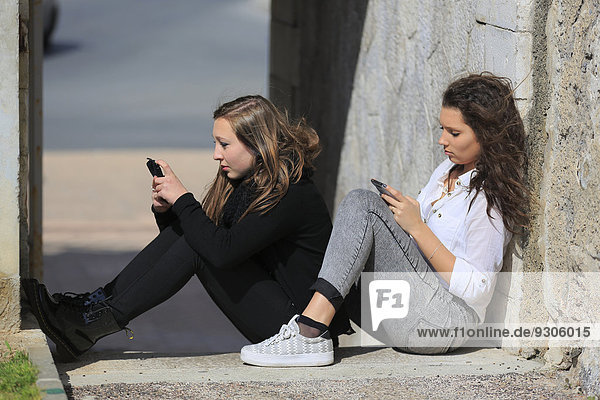 Zwei Freundinnen  Teenager  sitzen auf dem Boden vor einer Mauer und schreiben SMS auf ihren Handys  Menton  Provence-Alpes-Côte d?Azur  Frankreich