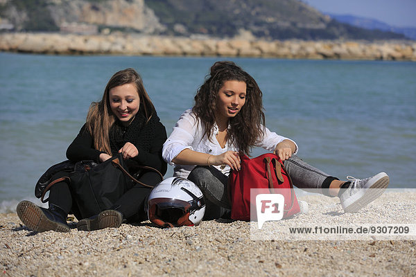 Zwei Freundinnen  Teenager  sitzen am Strand mit Taschen und Sturzhelm  Menton  Alpes-Maritimes  Provence-Alpes-Côte d'Azur  Frankreich