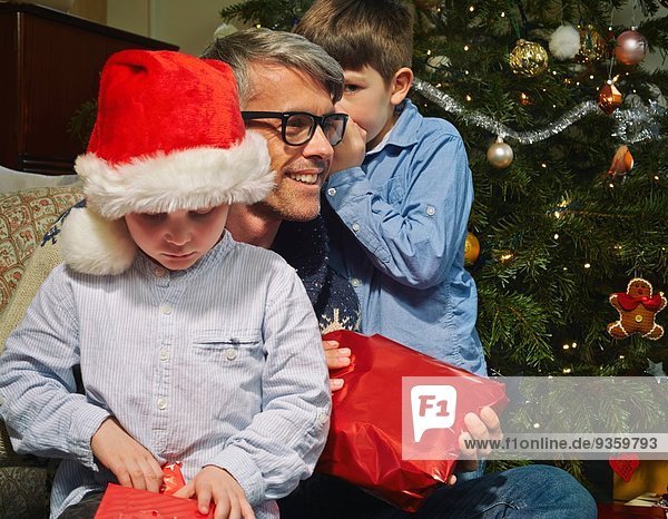 Junge flüstert dem Vater zu  während der Bruder das Weihnachtsgeschenk öffnet.