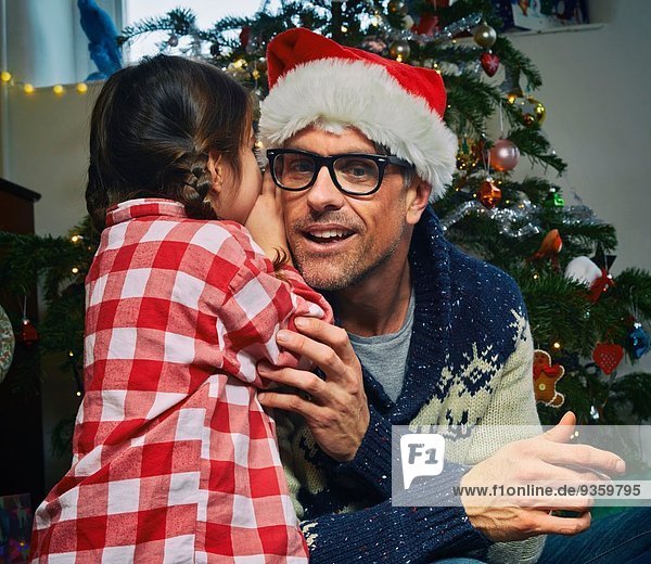 Tochter flüstert Vater zu Weihnachten mit Weihnachtsmütze zu