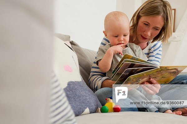 Mature mother and baby daughter on sitting room sofa reading storybook