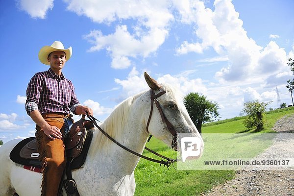 Porträt eines jungen Mannes in Cowboy-Ausrüstung zu Pferd auf der Landstraße