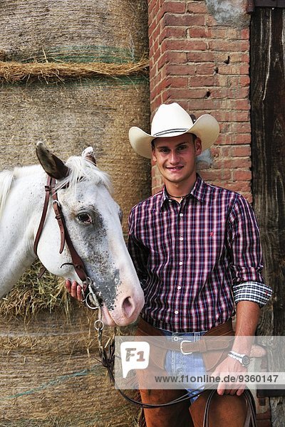 Portrait eines jungen Mannes in Cowboy-Ausrüstung mit Pferd im Außenstall