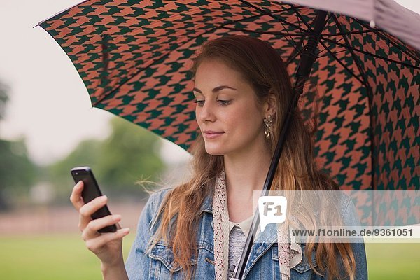 Junge Frau mit Regenschirm SMS auf Smartphone im Park
