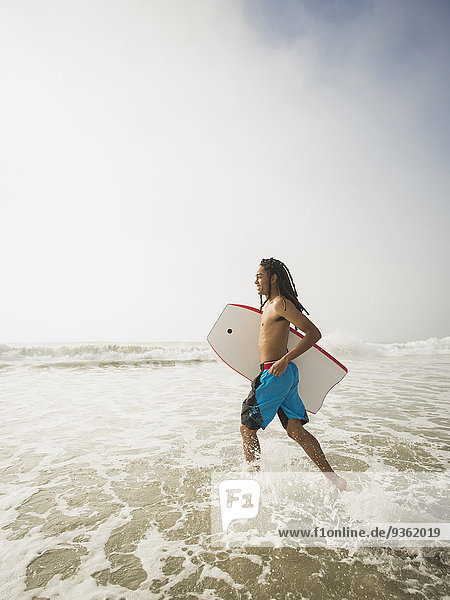 Black teenage boy carrying boogie board in waves