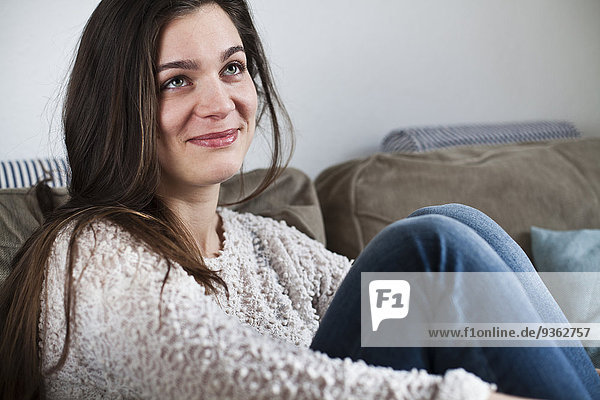 Porträt einer lächelnden jungen Frau auf der Couch in ihrem Wohnzimmer