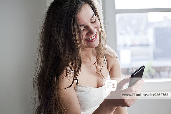 Porträt einer lächelnden jungen Frau mit ihrem Smartphone zu Hause