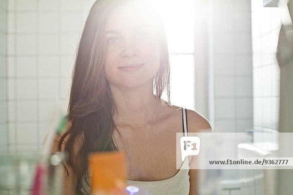 Porträt einer lächelnden jungen Frau  die ihr Spiegelbild im Badezimmer betrachtet.