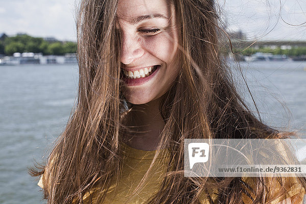 Deutschland  Köln  Porträt einer lächelnden jungen Frau mit wehendem Haar vor dem Rhein stehend