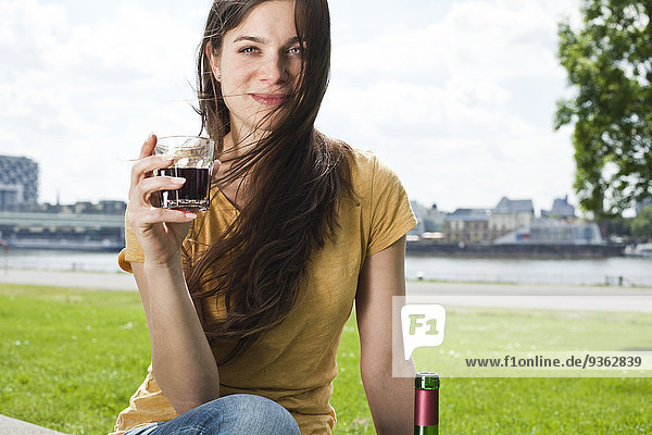 Germany  Cologne  portrait of smiling young woman with glass of red wine sitting in front of Rhine River