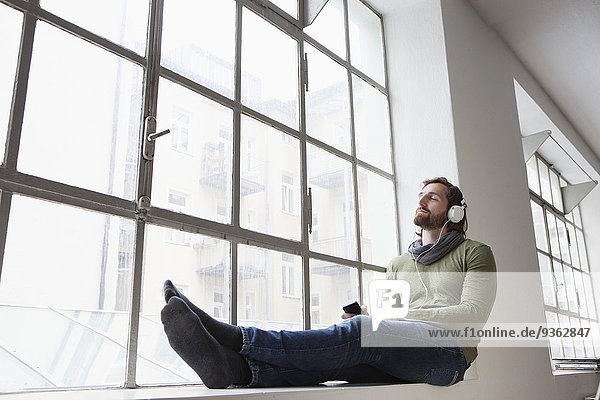 Porträt eines jungen Mannes  der auf einer Fensterbank in einem Büro sitzt und mit Kopfhörern Musik hört.