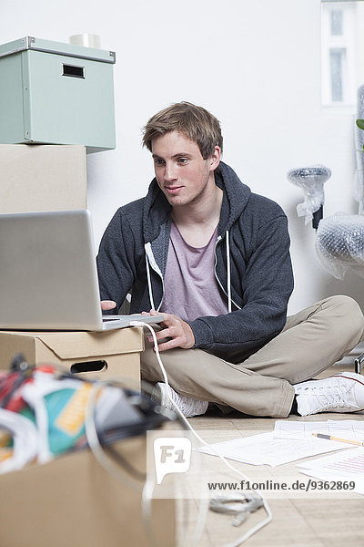 Portrait of young man sitting on ground between cardboard boxes in an office using his notebook