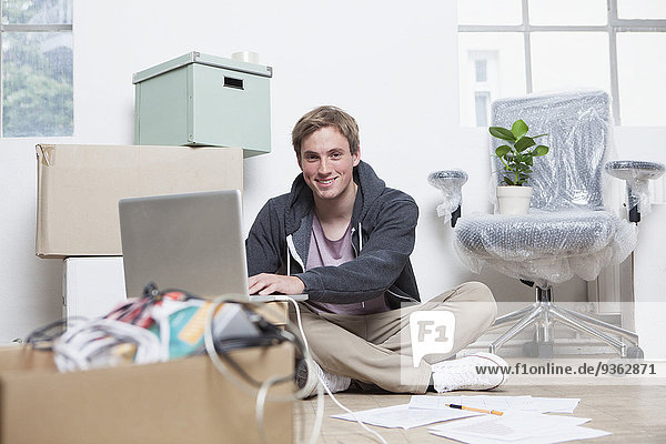 Porträt eines jungen Mannes  der mit seinem Notizbuch zwischen Pappkartons in einem Büro sitzt.