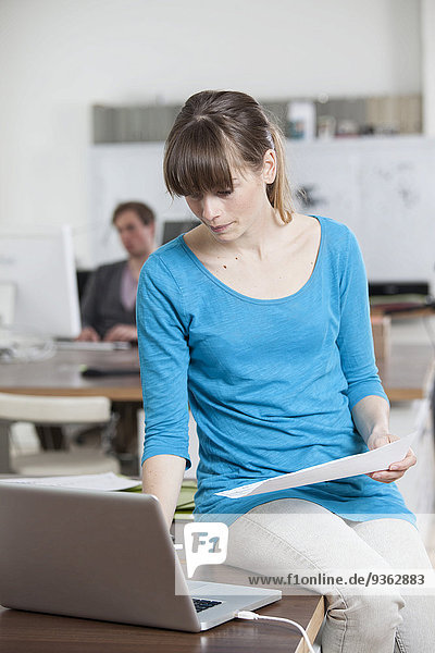 Junge Frau sitzt auf ihrem Schreibtisch in einem Büro mit Laptop