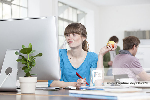 Porträt einer jungen Frau  die einen Apfel an ihrem Schreibtisch in einem Kreativbüro isst.