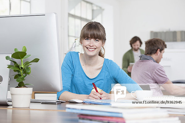 Porträt einer lächelnden jungen Frau  die an ihrem Schreibtisch in einem kreativen Büro sitzt.