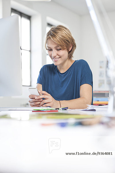 Porträt einer lächelnden jungen Frau mit Smartphone am Schreibtisch in einem Kreativbüro