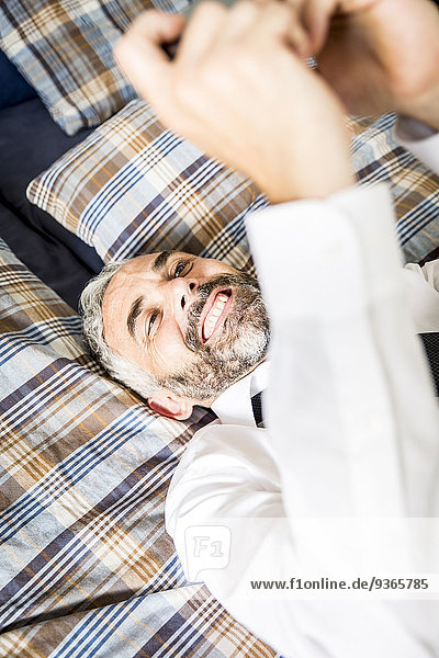 Porträt eines glücklichen Geschäftsmannes  der auf seinem Bett liegt und auf sein Smartphone schaut.
