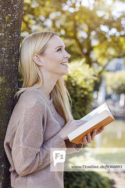 Lächelnde junge Frau mit geöffnetem Buch am Baumstamm lehnend