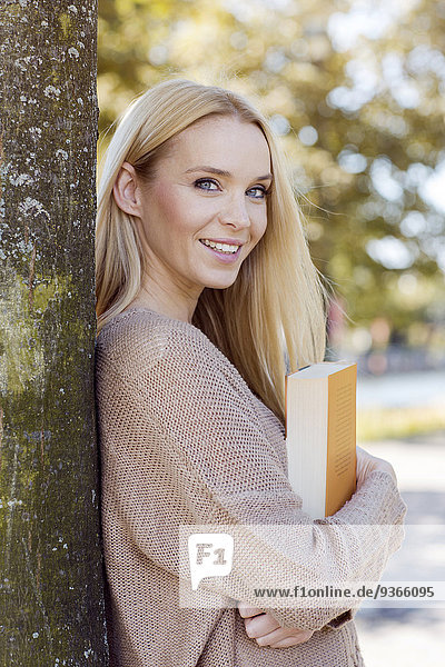 Porträt einer lächelnden jungen Frau mit Buch am Baumstamm lehnend