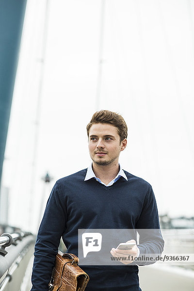 Deutschland  Hessen  Frankfurt  Portrait eines jungen Geschäftsmannes auf einer Brücke mit Aktentasche und Smartphone