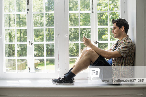 Am Fenster sitzender Mann mit einem Mobiltelefon in der Hand.