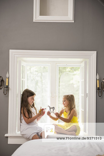 Zwei gemeinsam spielende Mädchen  die drinnen auf einem Fensterplatz sitzen.