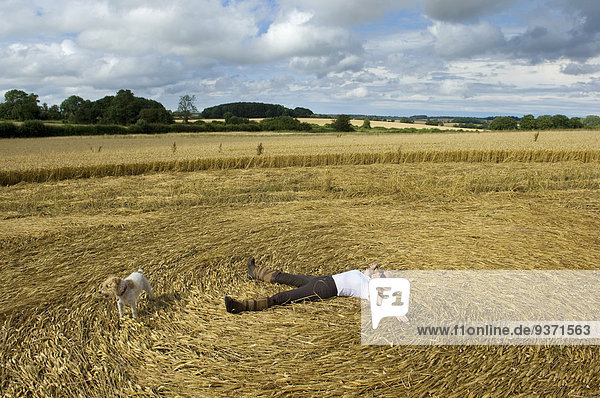 Ein Bauer  der auf dem Rücken in den Stoppeln eines frisch geschnittenen Getreidefeldes liegt  mit ausgebreiteten Armen und Beinen  wodurch ein Muster im Stroh entsteht.