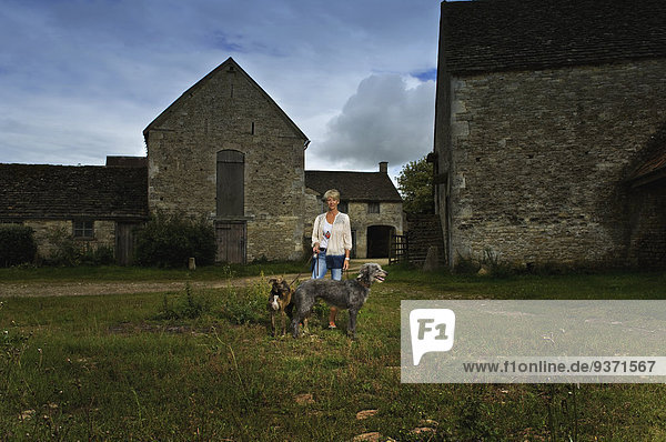 Eine Frau  die mit zwei großen Taumelhunden auf einem Bauernhof im Schatten steht.